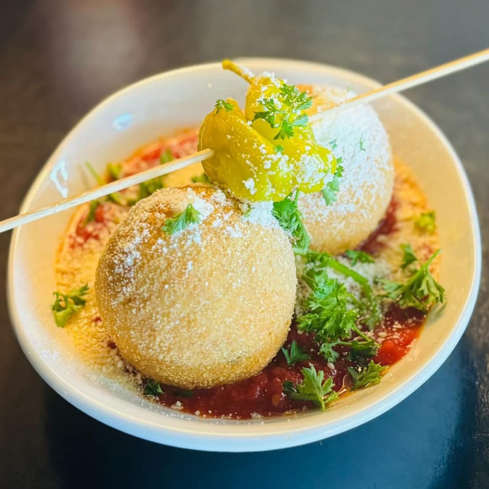 Two golden-brown arancini balls on skewers, garnished with peppers, parsley, and powdered sugar.