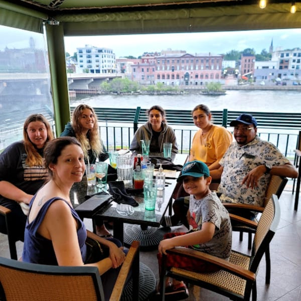 A family of seven smiles at a table on GS Steamers Bar & Grill's outdoor patio overlooking a river.