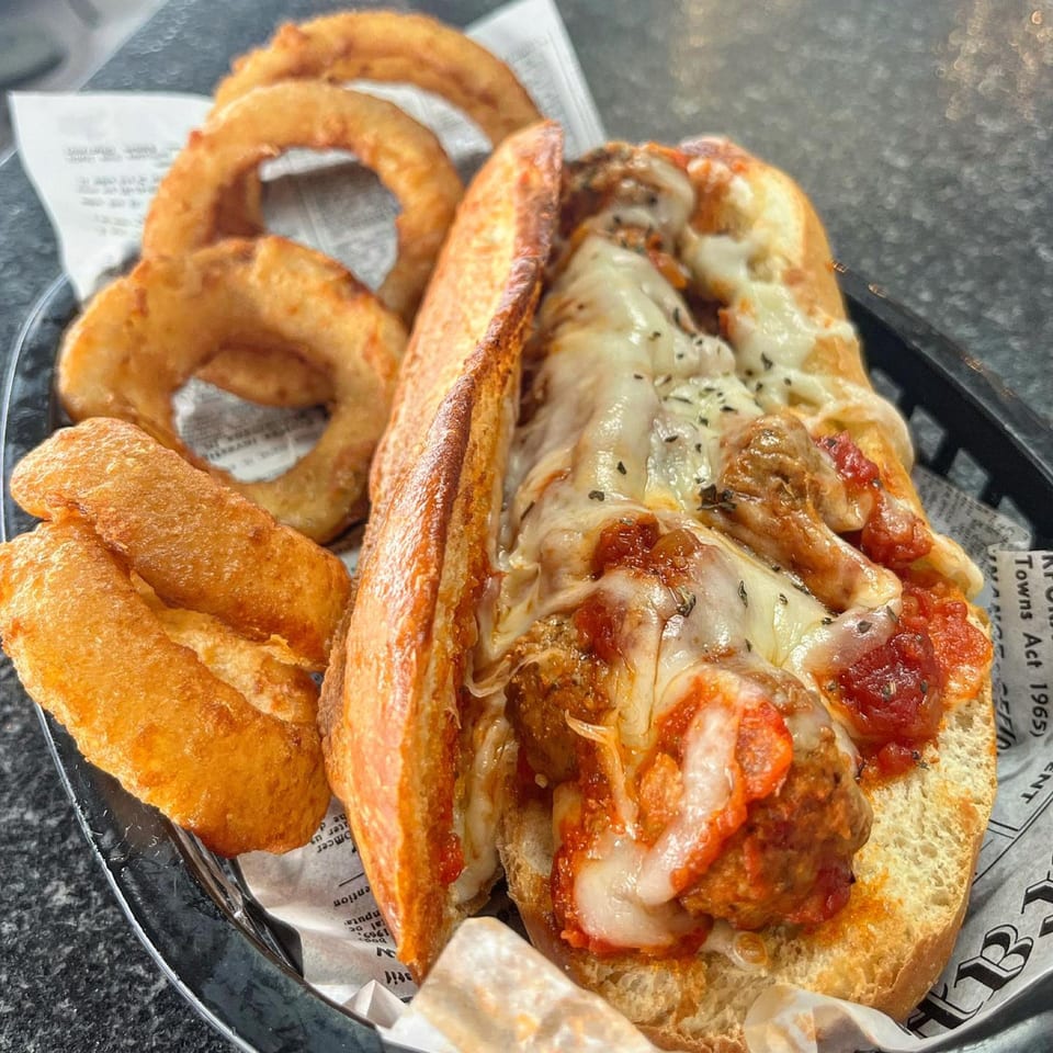 A meatball sub with melted cheese and marinara sauce, served with golden onion rings in a basket.