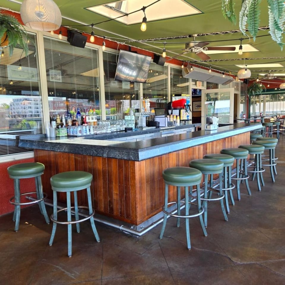 Long wooden bar with green stools, liquor bottles, and glasses, under a green ceiling with string lights.